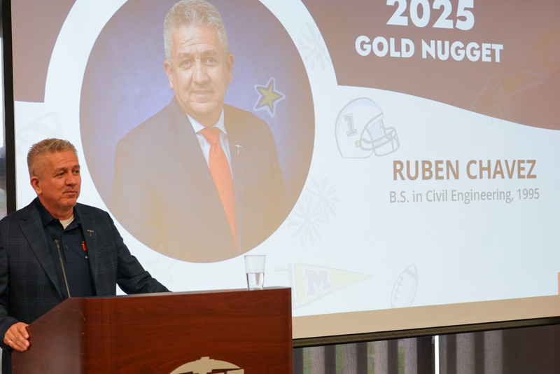 Individual stands at a podium delivering remarks during a UTEP Engineering homecoming gathering, with a large presentation slide recognizing the 2025 Gold Nugget award recipient Ruben Chavez and displaying academic credentials in Civil Engineering behind the speaker.
